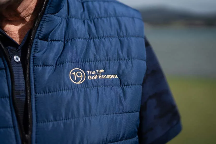 Close-up of a person's upper body wearing a blue quilted vest with the logo and text 'The 19th Golf Escapes' embroidered on it, outdoors near a golf course.