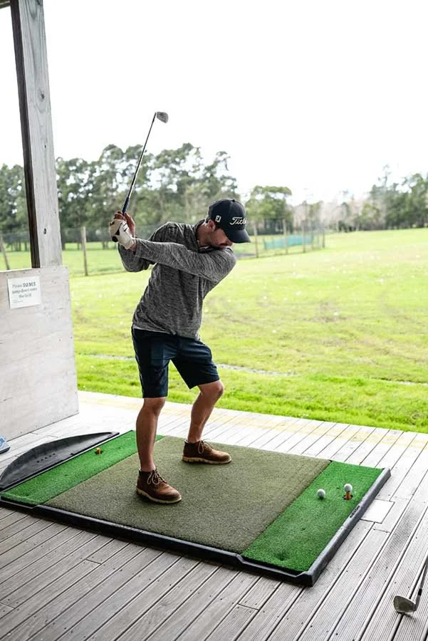 A young man practicing his golf swing at an indoor driving range, on a small artificial turf mat with outdoor grass and trees in the background.