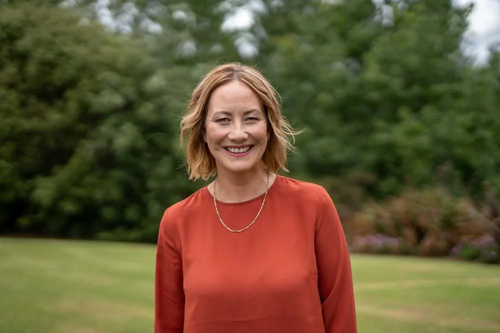 Woman with short, wavy blonde hair smiling outdoors in front of green trees, wearing a reddish-brown blouse and a delicate necklace.