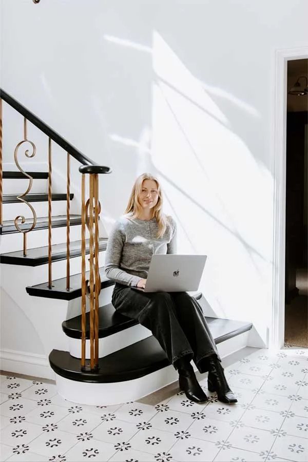 A woman sitting on a black and white curved staircase with a laptop, smiling in a bright room with white walls and patterned tile floor.