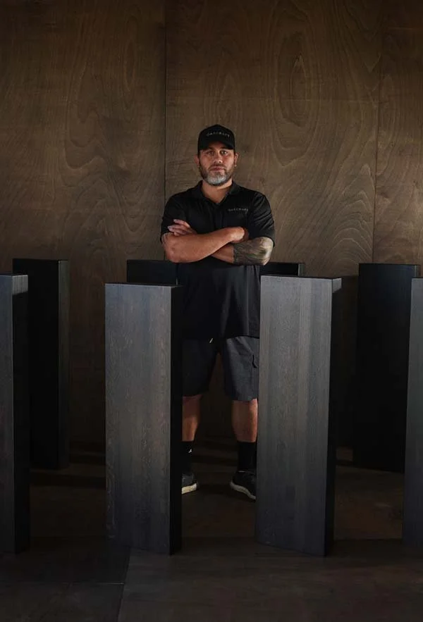 A man standing with arms crossed in front of a dark wooden wall, surrounded by black podiums, wearing a black cap, black shirt, shorts, and sneakers.