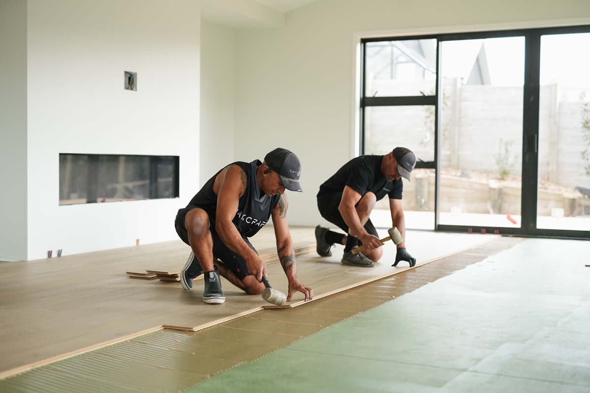 Two men installing hardwood flooring in a room with large windows.