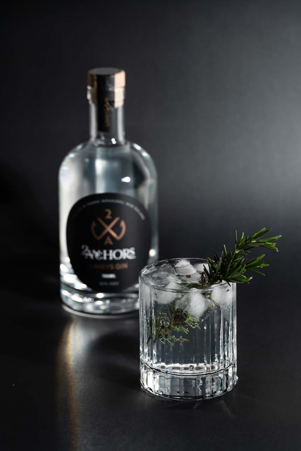 A bottle of Amplify Gin behind a glass of gin with ice and a sprig of rosemary garnish on a black surface and dark background.