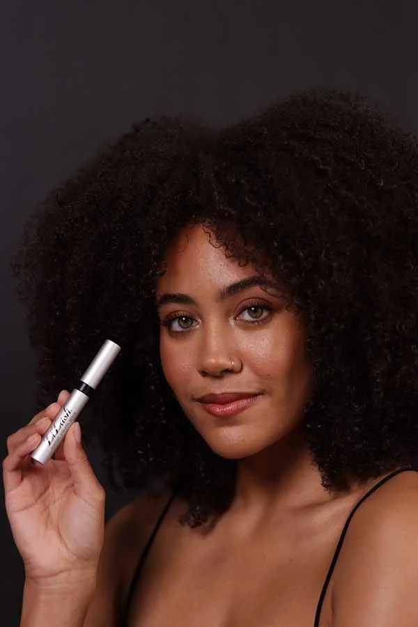 Woman with voluminous curly hair holding a white and gray cosmetic container, smiling at the camera against a dark background.