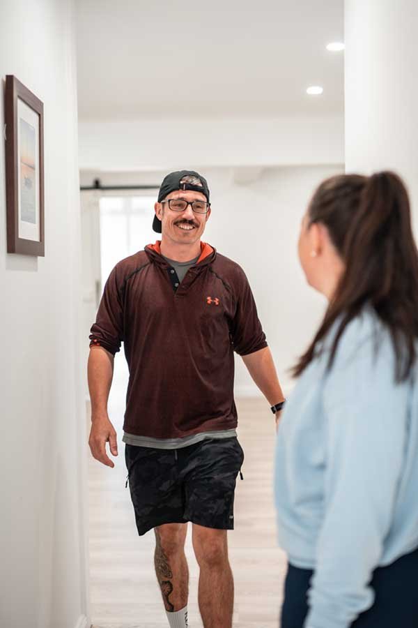 A man with glasses, a cowboy hat worn backwards, a goatee, and tattoos on his leg, smiling and walking in a hallway, engaging in conversation with a woman with long dark hair pulled back, wearing a light blue top.