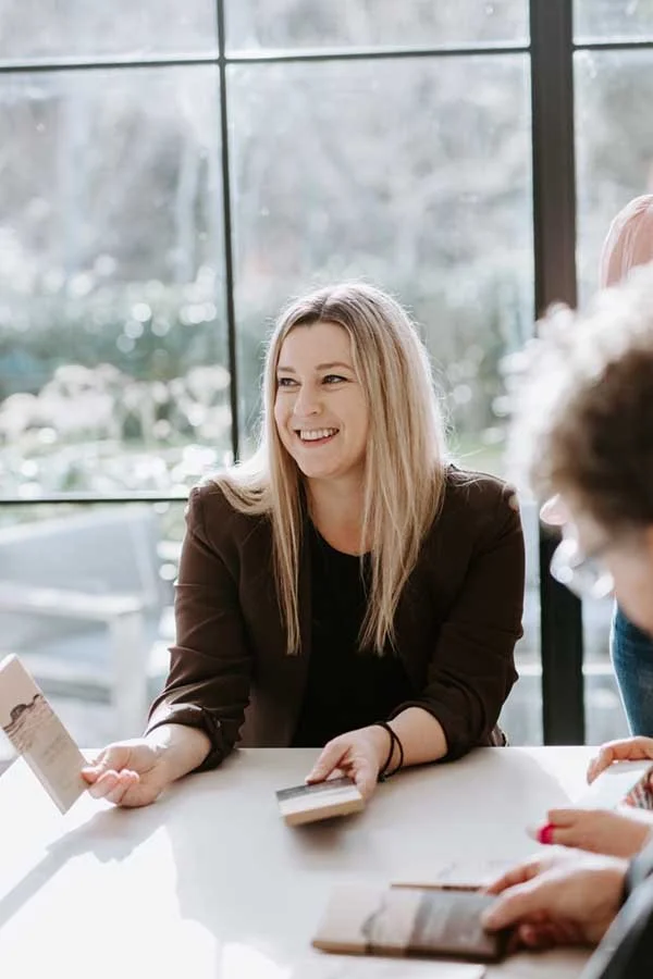 A woman with blonde hair smiling and holding a pamphlet or brochure in her right hand, seated at a table with others, in front of a large window with natural light.