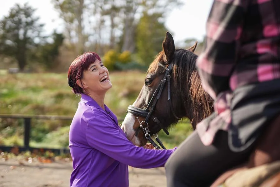 A woman with short reddish hair in a purple jacket smiling while hugging a brown horse with a black mane, outdoors with trees in the background.