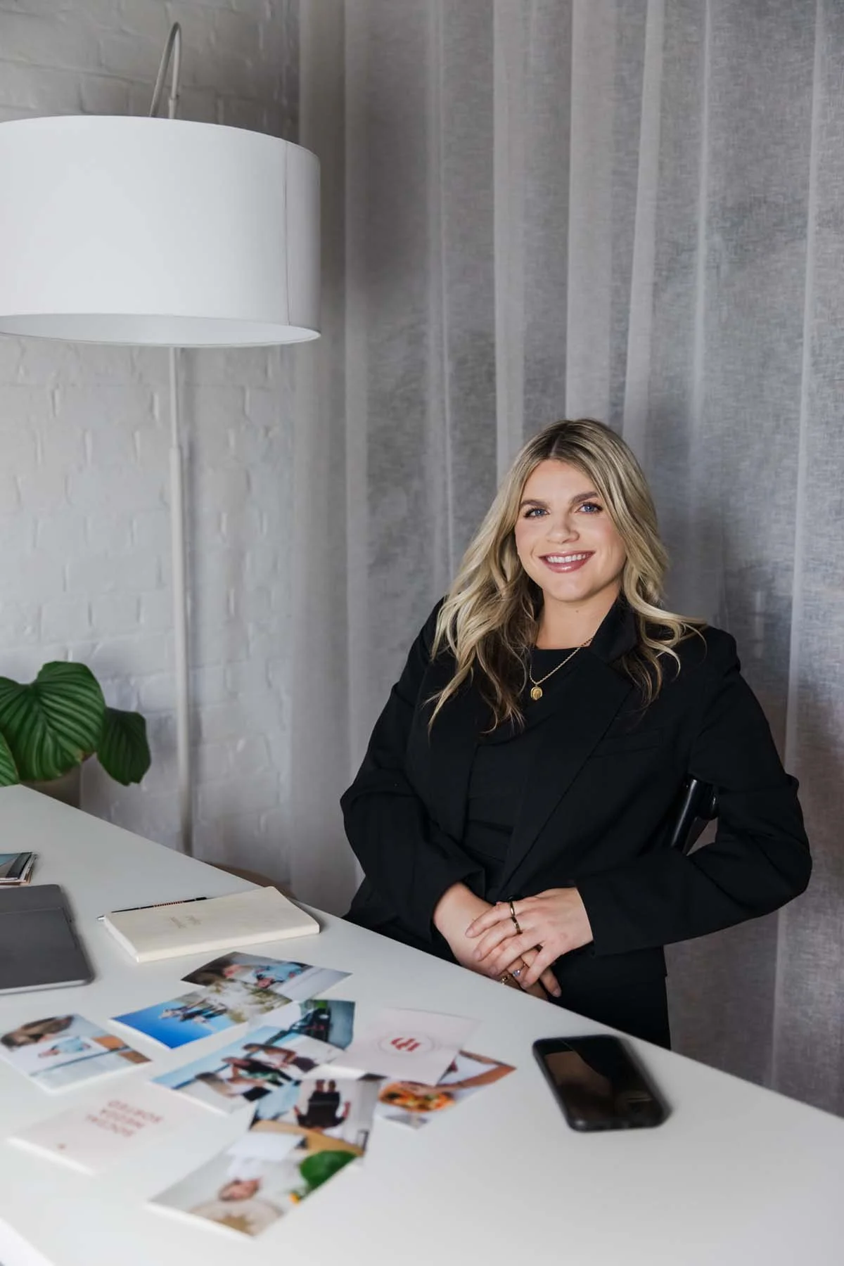 A woman with blonde hair smiling at a desk with photos, a smartphone, and a notebook in front of her, in a modern office with a white brick wall and sheer curtains.