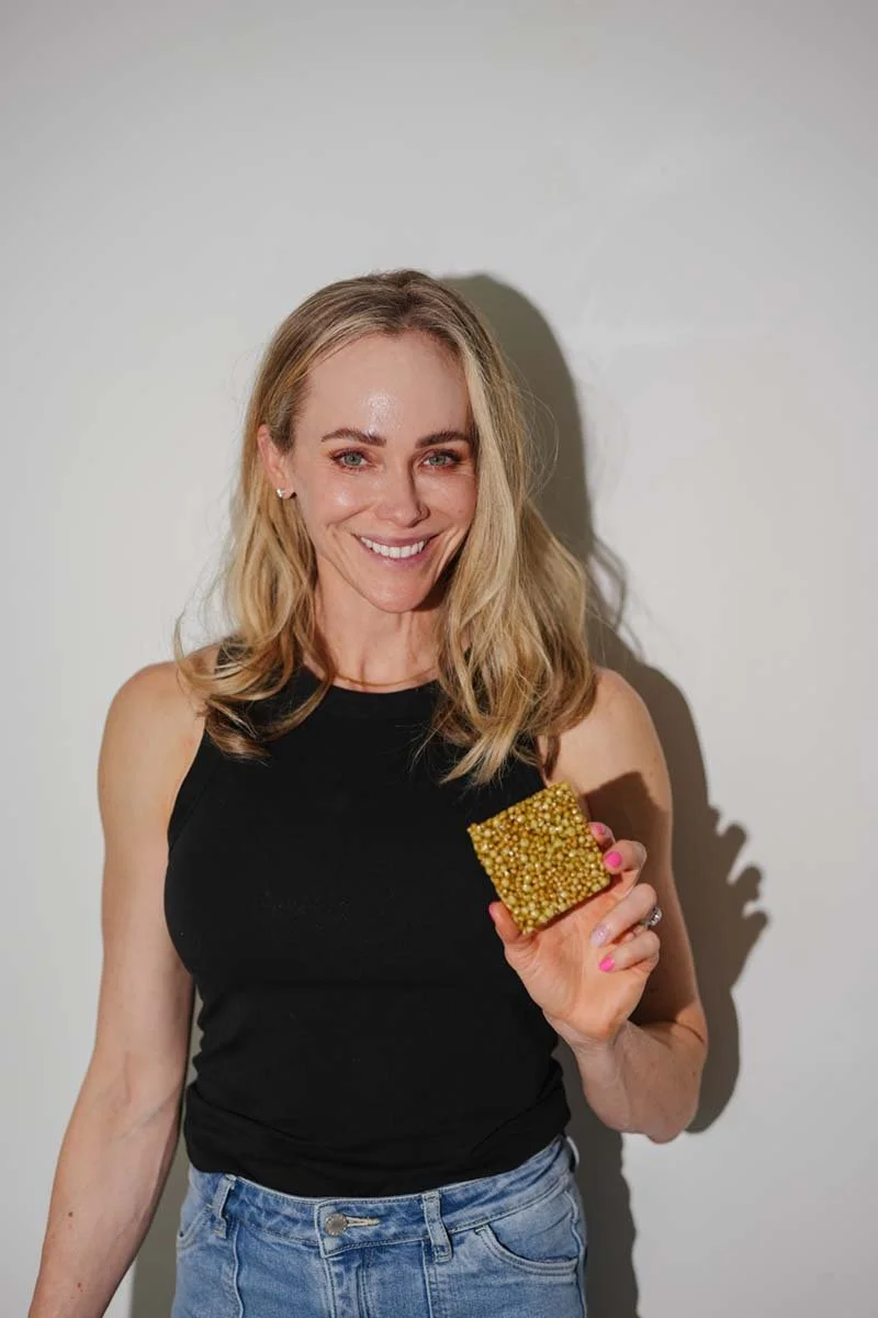 A woman with blonde hair smiling and holding a golden beaded square accessory or item, standing against a plain light wall.