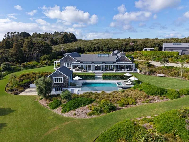 A large modern house with gray roof and white exterior, situated on a lush green landscape with a swimming pool in the backyard, surrounded by well-manicured lawn and landscaped garden, under a partly cloudy sky.