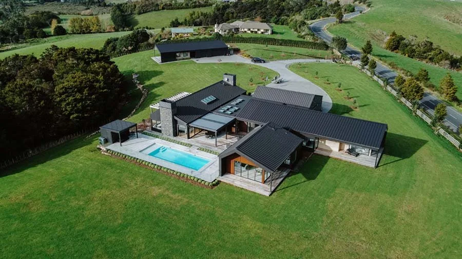 A modern house with a swimming pool, surrounded by a green lawn and trees, with a winding driveway leading to the property.