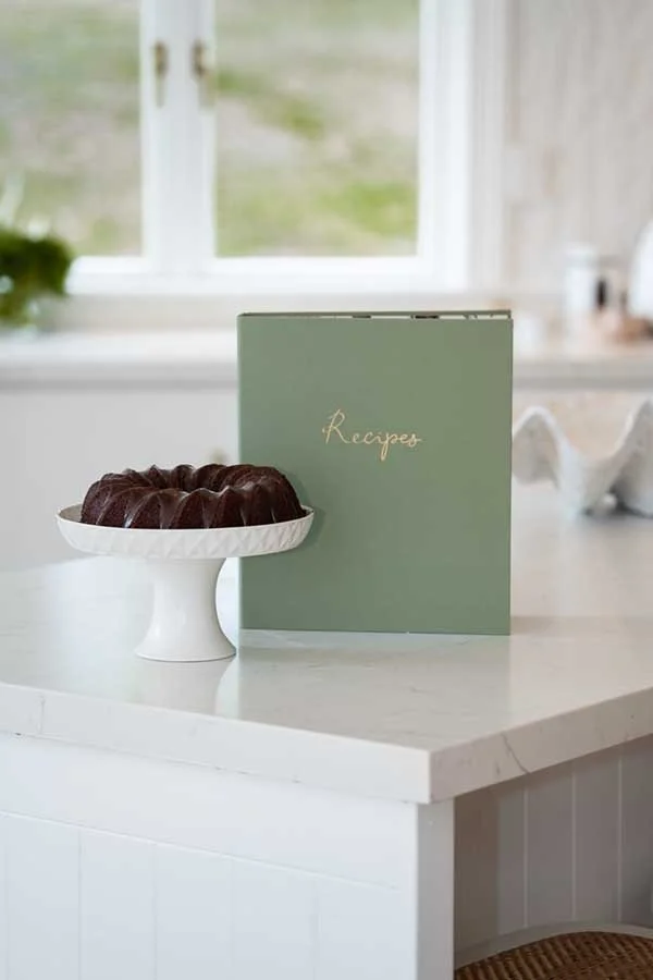 Chocolate bundt cake on a white cake stand with a green recipe book in the background on a white kitchen counter.