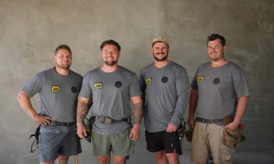 Four men standing together smiling, wearing grey t-shirts, against a neutral background.