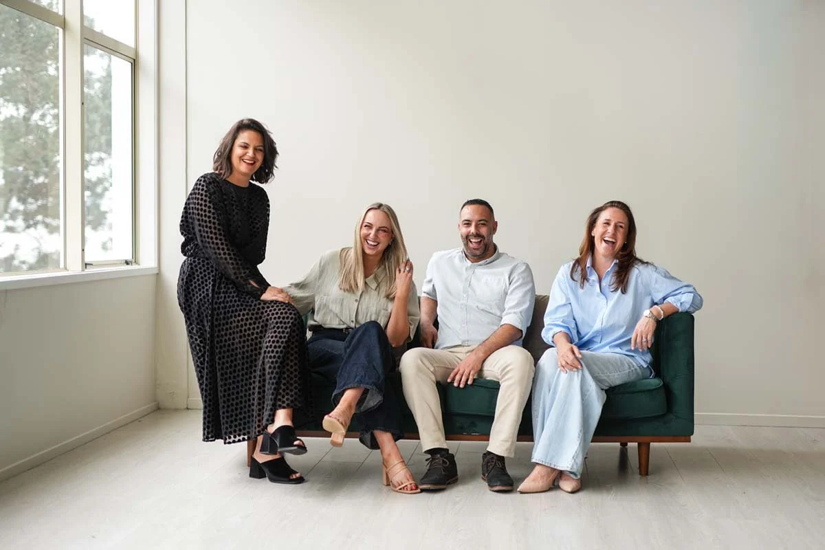 Four people sitting and standing on a green couch in a bright room, smiling and laughing, with a large window to the left.