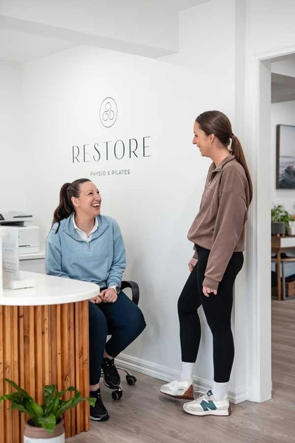 Two women smiling and talking in a wellness or fitness clinic with a sign that says "Restore Physio & Pilates" on the wall.