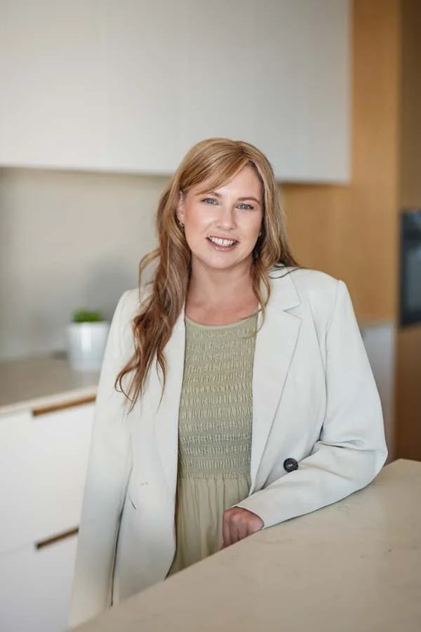 A woman with long red hair smiling and wearing a light green dress and white blazer, standing in a modern kitchen.