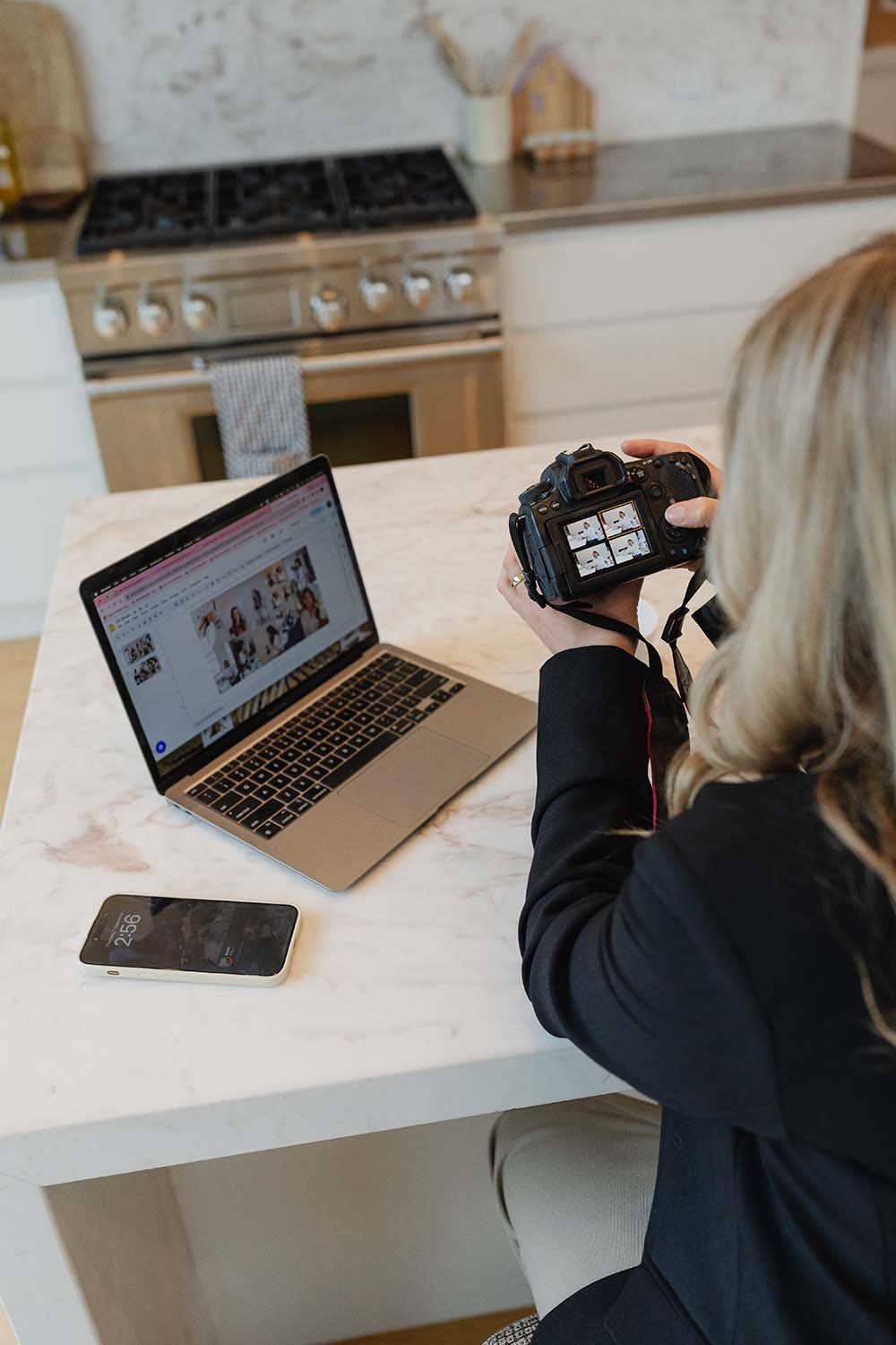 A woman with blonde hair sitting at a kitchen table, holding a camera and taking a photo of her laptop screen. The laptop displays a collage of photos or a website with multiple images. A smartphone is placed on the table beside the laptop. The kitchen has a stove, oven, and a white countertop with abstract wall art in the background.