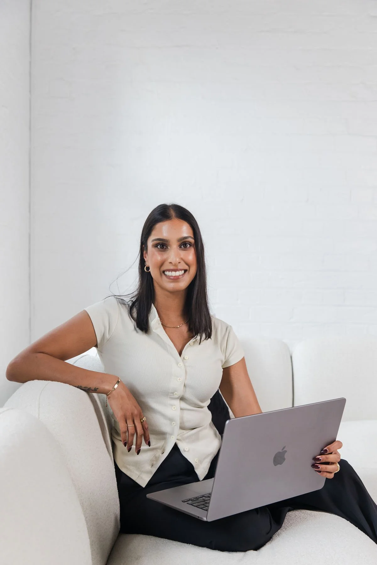 A woman with dark hair, wearing a white short-sleeve blouse, sitting on a light-colored sofa, smiling, holding a silver MacBook laptop, against a white brick wall background.
