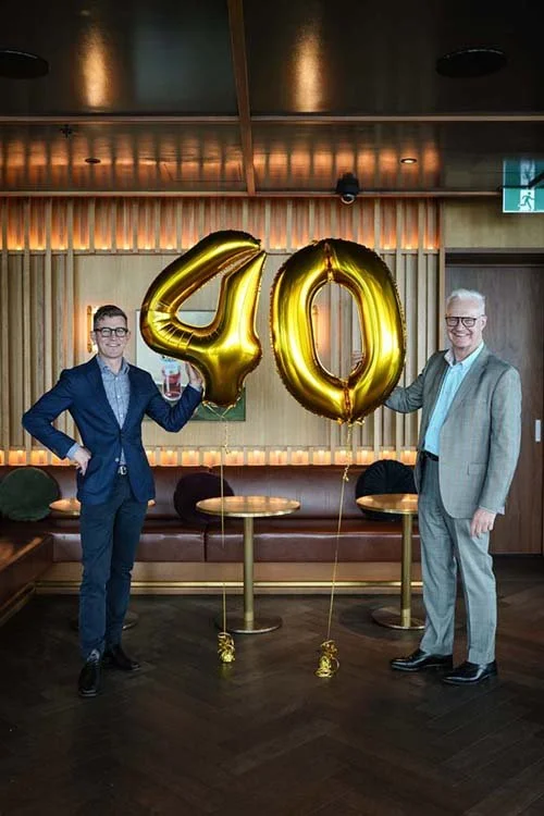 Two men in suits holding large gold balloons shaped as the number 40 in a modern room with wooden paneling and seating.