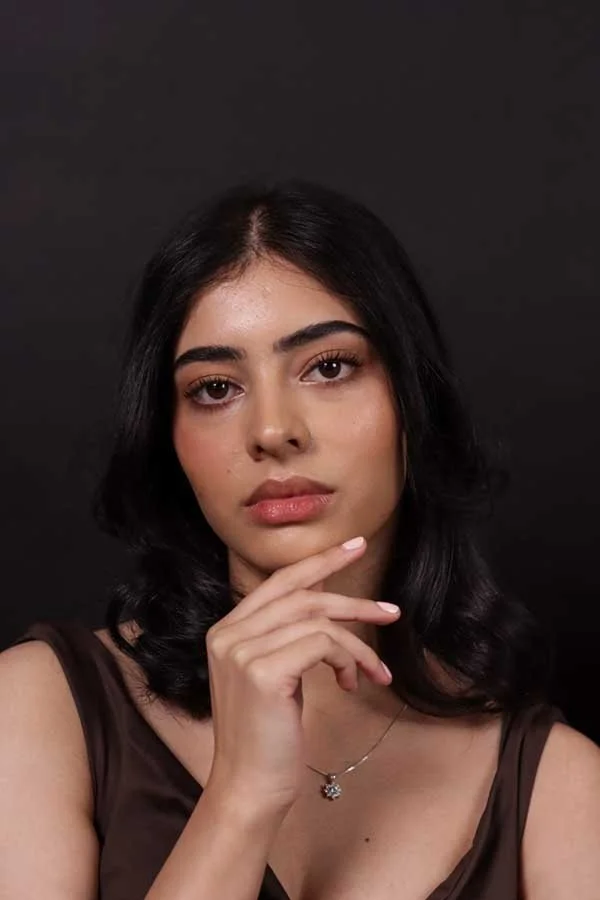 A young woman with dark hair and a neutral expression, wearing a sleeveless brown top and a delicate necklace, posing against a dark background.
