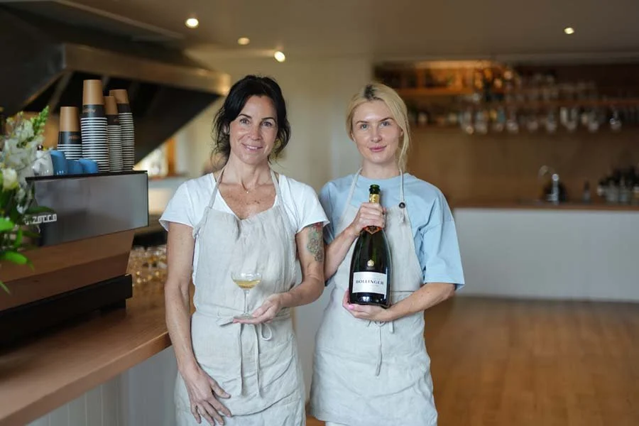 Two women in aprons stand in a bar or restaurant, one holding a glass of wine and the other holding a bottle of sparkling wine or champagne.
