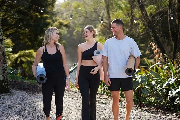 Three people walk together outdoors carrying yoga mats, smiling and chatting on a trail in a lush, green park.