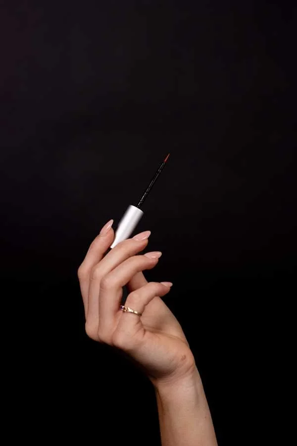 A hand with a gold ring on the ring finger holding a white container with an applicator brush, likely a nail or eyelash polish, against a black background.