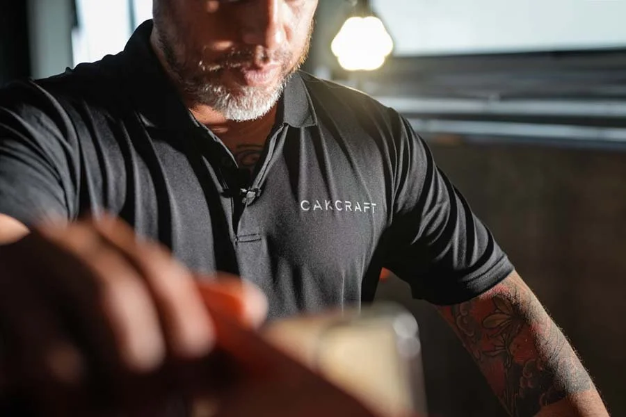 A man with a beard wearing a black polo shirt with the logo 'CAKCRAFT' on it, reaching towards the camera, with a tattooed arm and a blurred background.