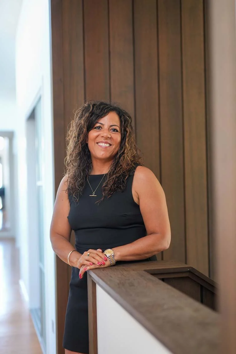 A woman with curly brown hair smiling and wearing a black sleeveless dress, standing indoors next to a wooden railing with a wood-paneled wall behind her.