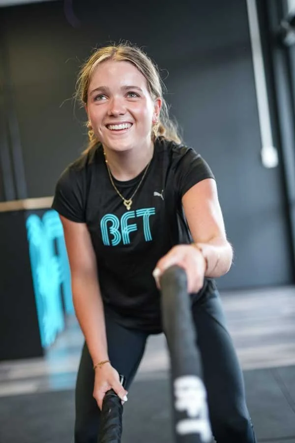 Young woman with blonde hair smiling while exercising with battle ropes in a gym.