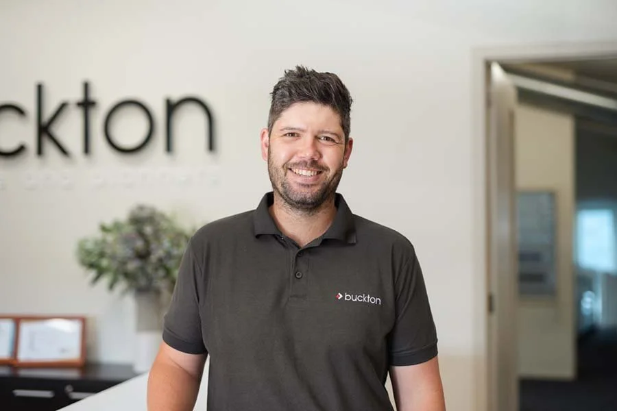 A man smiling in an office environment, wearing a black polo shirt with the 'buckton' logo. Behind him is a blurred logo on the wall, a potted plant, and a doorway leading to another room.