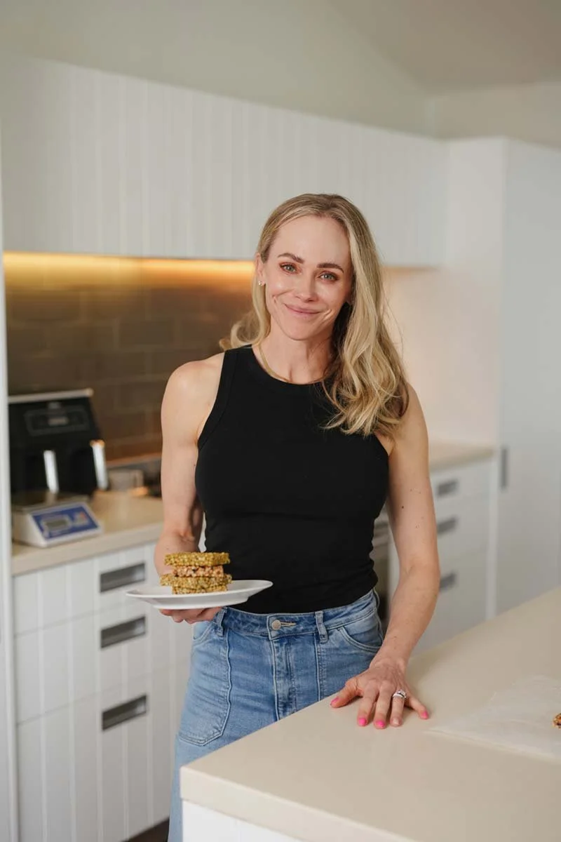 A woman with blonde hair, wearing a black sleeveless top and blue jeans, smiling and holding a plate of granola bars in a kitchen.