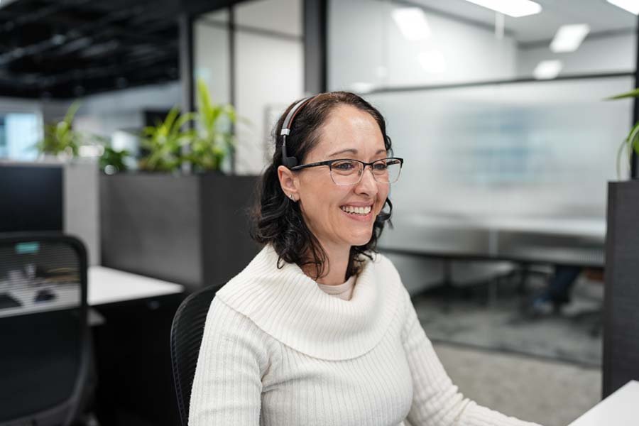 A woman with glasses and dark wavy hair working at a computer in an office, wearing a white sweater and a headset, smiling.