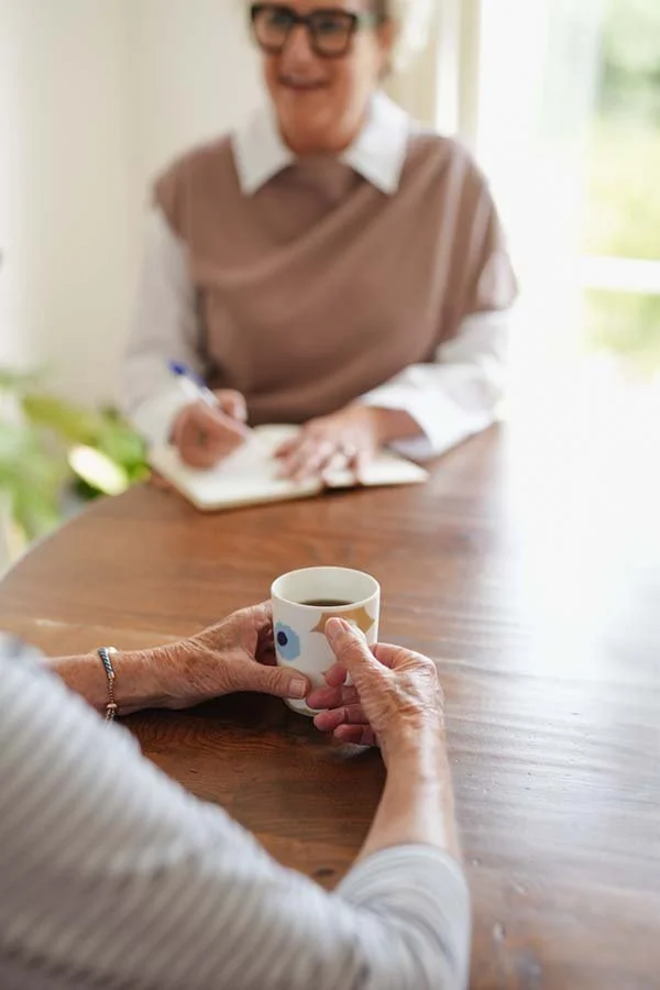 A person holding a coffee mug at a wooden table, with another person in the background taking notes.
