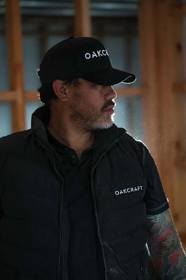 Man in black Oakley cap and Oakcraft vest, with tattoos on his arm, looking to the side indoors.