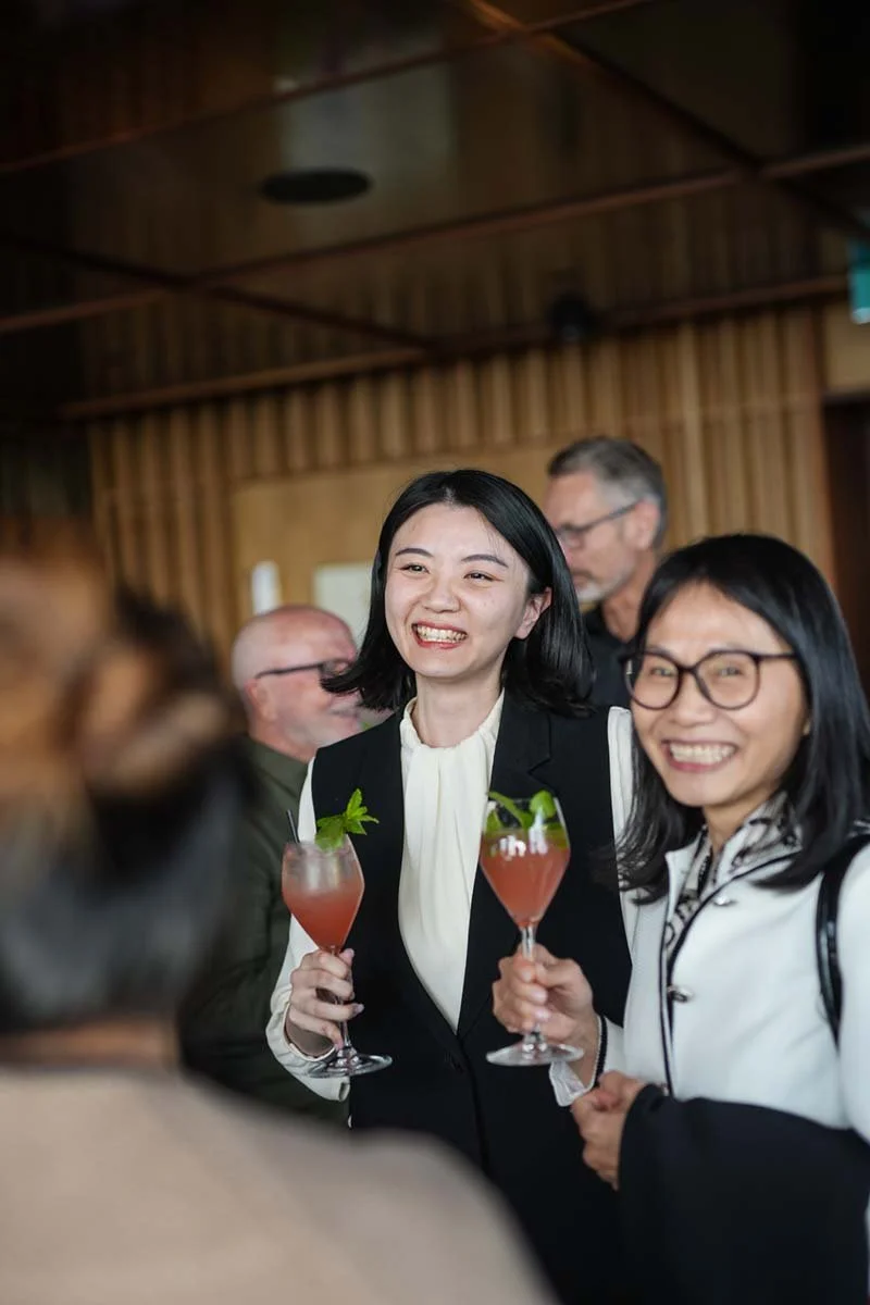Two women smiling and holding pink cocktails with mint leaves at a social event, with others in the background.