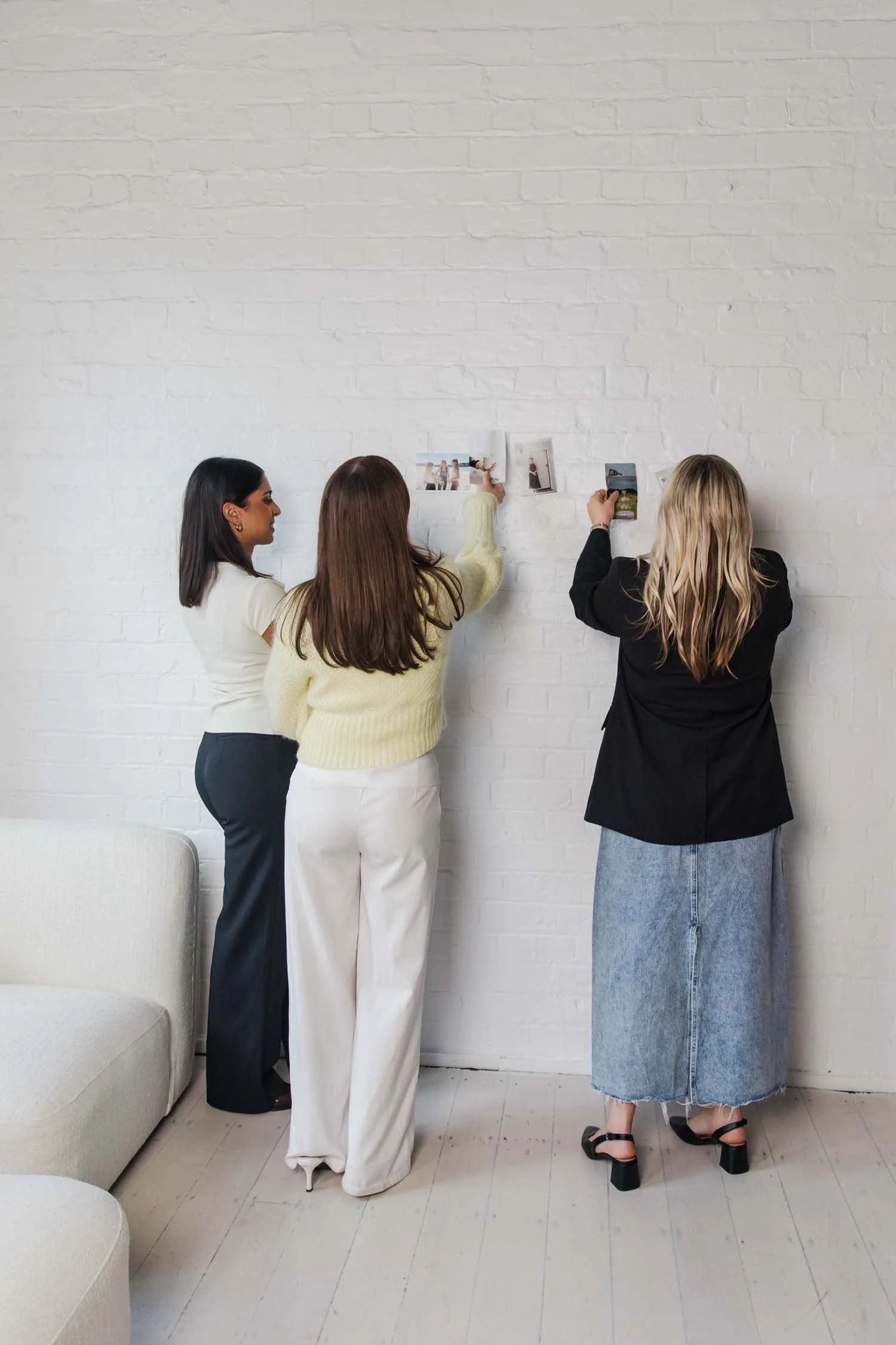 Three women hanging up photographs on a white brick wall in a room with light-colored wooden flooring and a beige sofa.