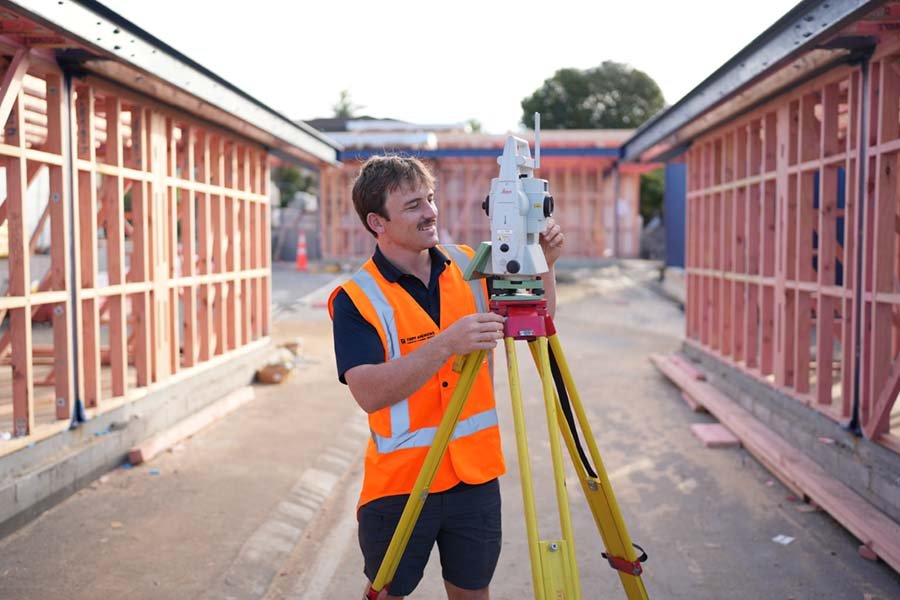 A construction worker using a leveling instrument on a construction site with wooden framing in the background.