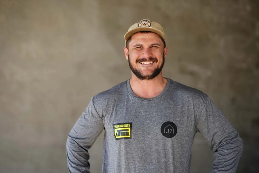 Smiling man wearing a beige cap, gray long-sleeve shirt with patches, and standing against a blurred neutral background.