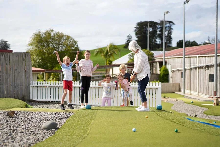 A group of children and an elderly woman playing mini golf outdoors on a cloudy day.