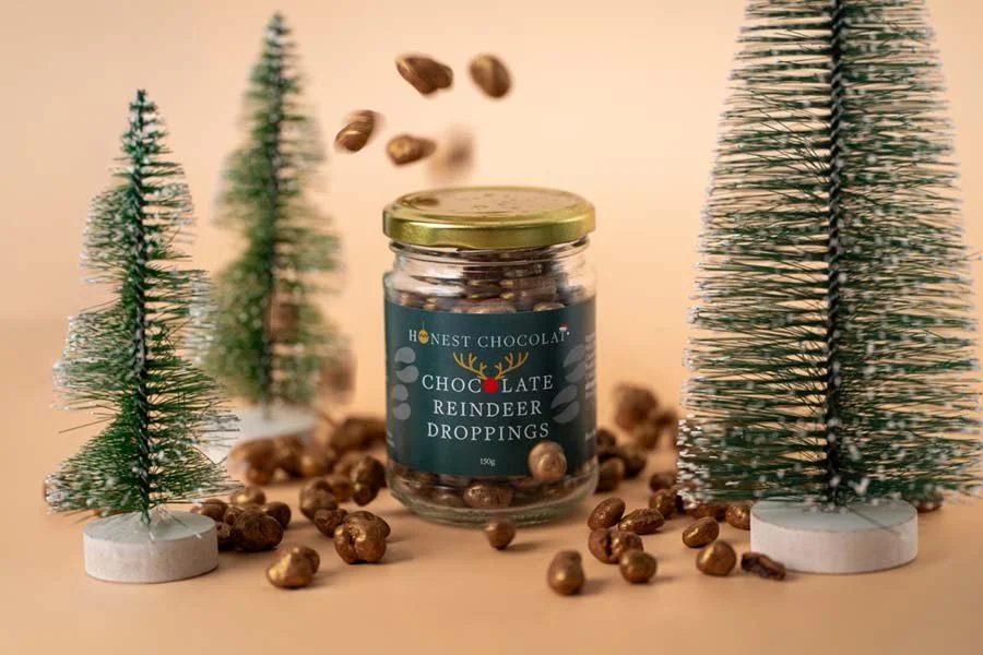 A jar labeled 'Honest Chocolat Chocolate Reindeer Droppings' surrounded by small decorative Christmas trees with some chocolate or candy pieces falling above the jar.