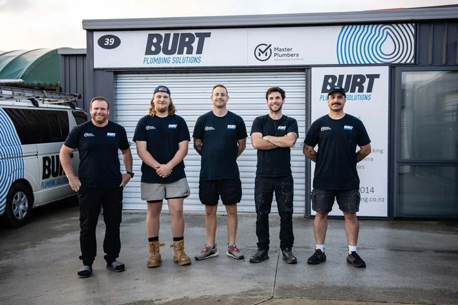 Five men standing in front of a Burton Plumbing Solutions building, wearing matching black company T-shirts, posing for a photograph.