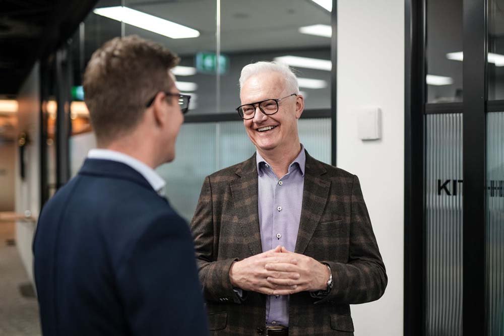 Two men talking in office, one smiling and wearing glasses and a plaid blazer, the other back facing camera wearing a dark suit and glasses.