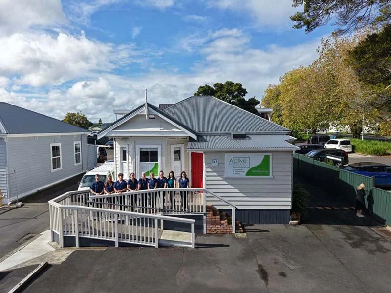 Group of people standing on a ramp in front of a small white building with a gray roof, parking lot, and trees in the background.