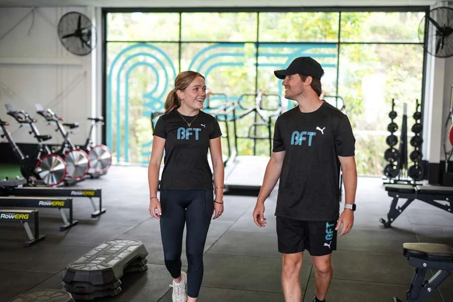 A man and a woman walking and talking inside a gym, both wearing black sports shirts with a logo that says BFT and Puma.