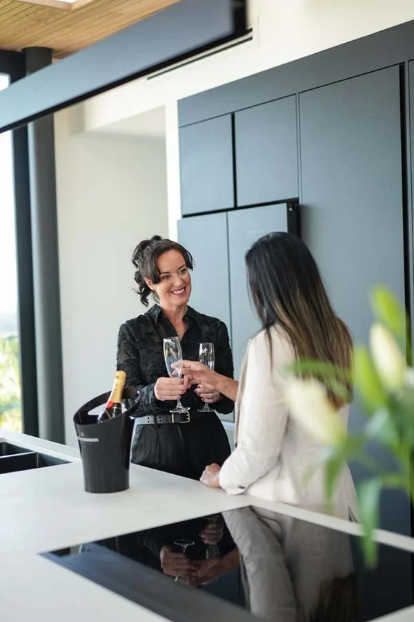 Two women chatting at a modern kitchen bar, toasting with champagne glasses, with a champagne bottle in an ice bucket on the counter.