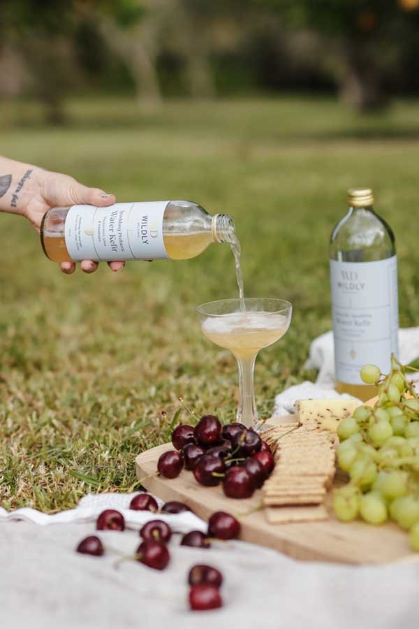 Person pouring white wine into a coupe glass on a picnic blanket with grapes, cheese, and crackers, outdoors in a park.