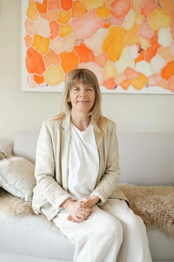 A woman with blonde hair sitting on a white sofa in front of an abstract painting with orange, yellow, and pink shapes.