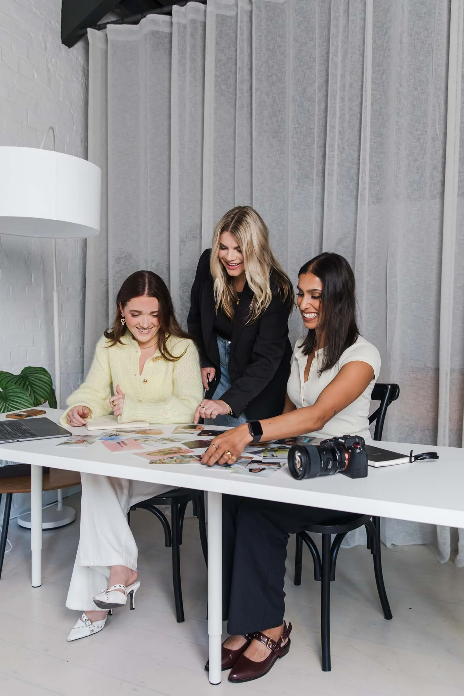 Three women collaborating at a white table, looking at photos and notes, with a laptop, camera, and notebook present in a well-lit modern office with gray curtains and a white brick wall.