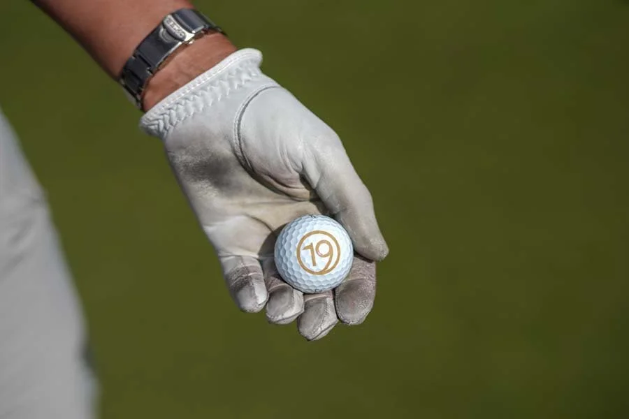 Person wearing a white glove holding a golf ball with the number 19 on it, on a green golf course.